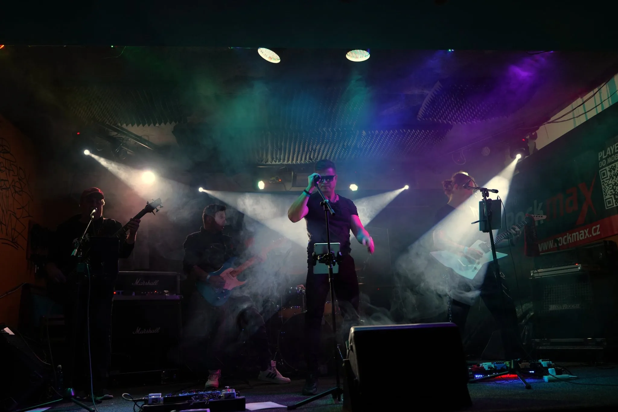 Přední řada kapely v kouři a barevných reflektorech v klubu Rockmax
